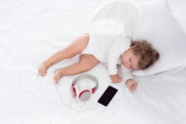 Маленький ангел с крыльями лежал на кровати с наушниками и смартфоном — стоковое фото