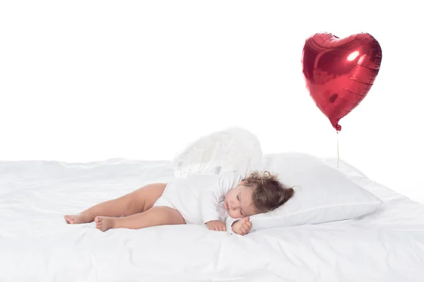 Маленький херувим с крыльями, спящий на кровати с сердечным шаром, изолированный на белом — стоковое фото