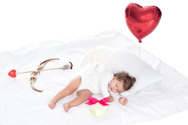 Little cherub with wings lying on bed with heart balloon, present, bow and arrow, isolated on white — Stock Photo