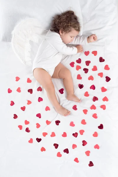 Cupidon avec des ailes dormant sur le lit avec des cœurs rouges — Photo de stock