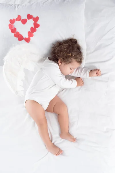 Adorable Cupidon avec des ailes dormant sur le lit avec des cœurs rouges — Photo de stock