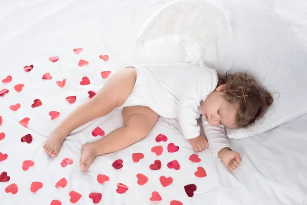 Little cupid with wings sleeping on bed with red hearts — Stock Photo