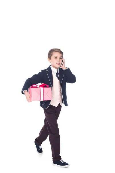 Niño sonriente hablando en el teléfono inteligente y sosteniendo la caja de regalo mientras camina aislado en blanco - foto de stock