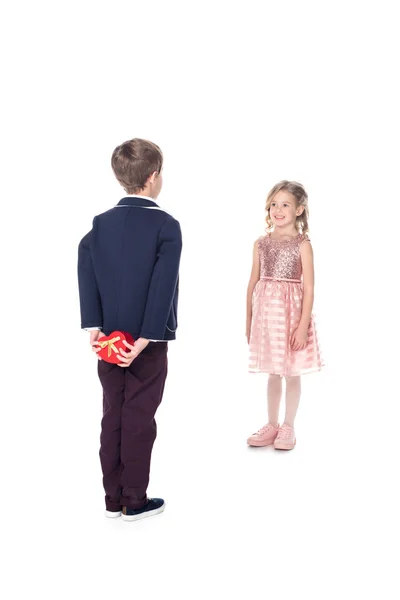 Вид сзади стильного мальчика в костюме, держащего подарочную коробку в форме сердца и смотрящего на улыбающуюся маленькую девочку в розовом платье, изолированном на белом — стоковое фото