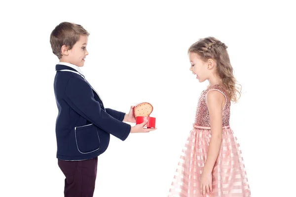 Вид збоку стильного хлопчика, що відкриває і представляє подарункову коробку у формі серця, щоб здивувати маленьку дівчинку ізольовану на білому — стокове фото