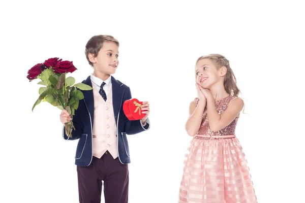 Schönes glückliches kleines Mädchen, das Junge mit rosafarbenen Blumen und herzförmigem Geschenkkarton ansieht, isoliert auf weiß — Stockfoto