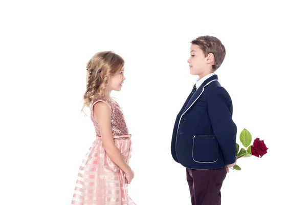 Вид сбоку на модного мальчика в костюме, держащего розовый цветок и смотрящего на восхитительную маленькую девочку в розовом платье, изолированном на белом — стоковое фото