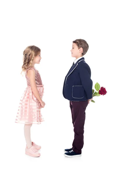 Vista lateral do menino elegante em terno segurando rosa flor e olhando para a menina bonita em vestido rosa isolado no branco — Fotografia de Stock