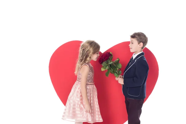 Vista lateral de niño pequeño presentando flores a hermosa niña y gran símbolo del corazón rojo detrás aislado en blanco - foto de stock