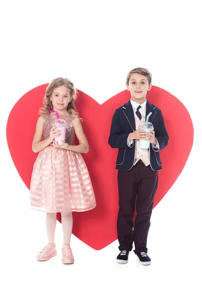 Adorables niños pequeños sosteniendo batidos en vasos de plástico y gran símbolo del corazón rojo detrás aislado en blanco - foto de stock