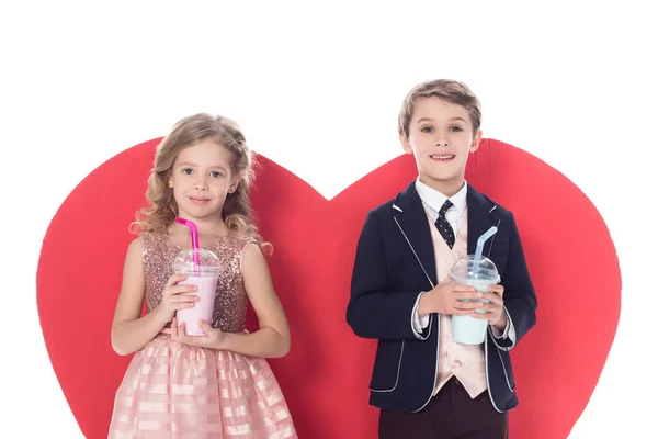 Lindos niños pequeños sosteniendo batidos en vasos de plástico y gran símbolo rojo del corazón detrás aislado en blanco - foto de stock