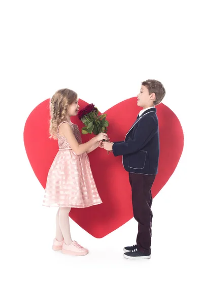 Vista lateral do menino bonito apresentando flores para adorável menina e grande símbolo coração vermelho atrás isolado no branco — Fotografia de Stock