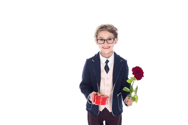 Entzückender kleiner Junge in Anzug und Brille mit rosafarbener Blume und herzförmiger roter Geschenkschachtel isoliert auf weiß — Stockfoto