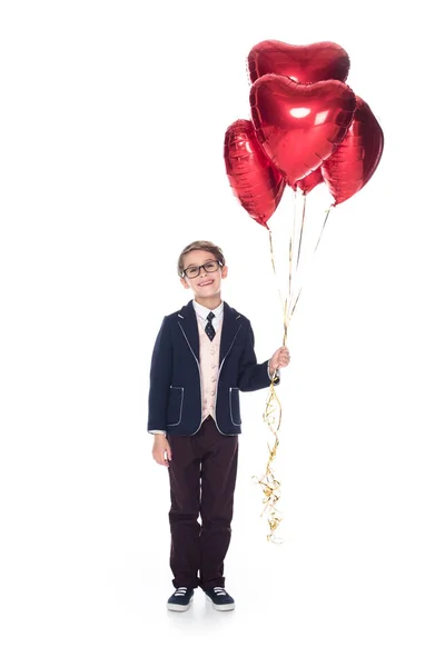 Cute little boy in suit and eyeglasses holding red heart shaped balloons and smiling at camera isolated on white — Stock Photo