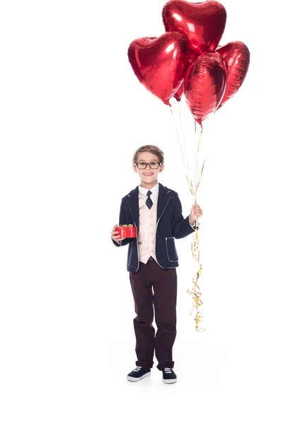 Adorable smiling little boy in suit and eyeglasses holding red heart shaped balloons and gift box isolated on white — Stock Photo