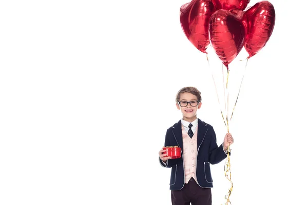 Menino bonito em terno e óculos segurando balões em forma de coração vermelho e caixa de presente isolado no branco — Fotografia de Stock