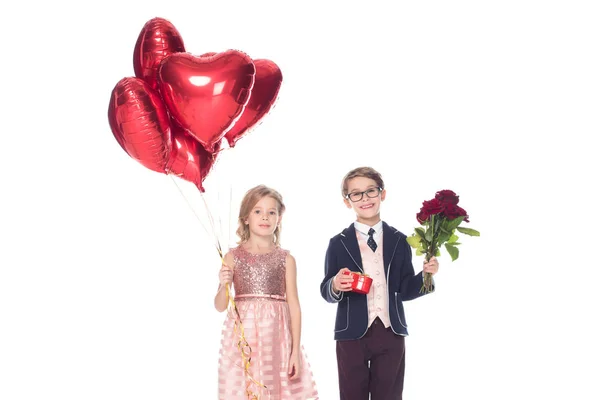 Adorable feliz pequeña pareja con globos en forma de corazón, caja de regalo y rosas sonriendo a la cámara aislada en blanco - foto de stock