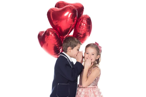 Stylish little boy holding heart shaped balloons and whispering to smiling beautiful little girl isolated on white — Stock Photo