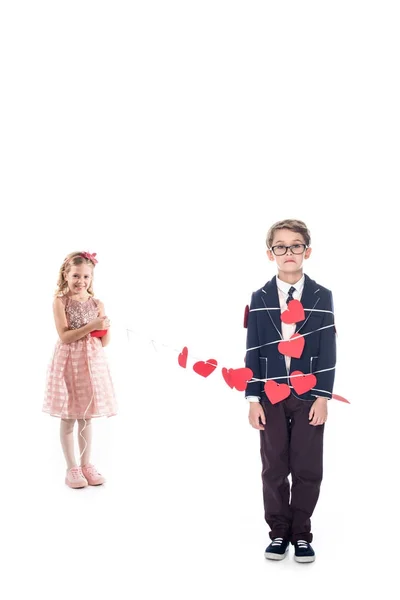 Adorable sonriente niña atando elegante chico con cuerda y rojo corazones aislado en blanco - foto de stock