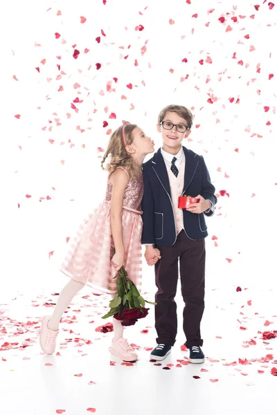 Netter kleiner Junge in Brille und Anzug mit Geschenkbox und lächelnd in die Kamera, während Mädchen mit Rosen ihn auf Weiß mit fallendem herzförmigem Konfetti küssen können — Stockfoto