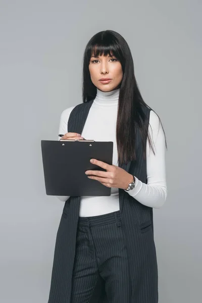 Businesswoman standing with clipboard and looking at camera isolated on gray — Stock Photo