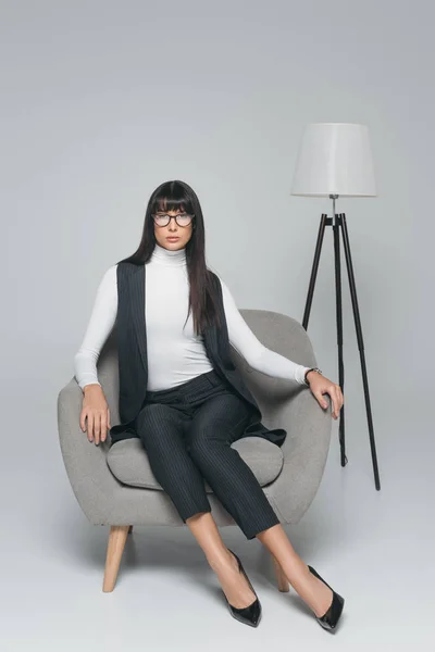 Attraktive brünette Frau sitzt im Sessel und schaut in die Kamera auf grau — Stockfoto