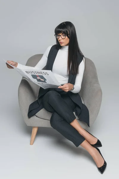 Attractive brunette businesswoman reading newspaper in armchair on gray — Stock Photo