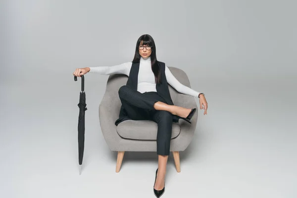 Красива брюнетка бізнес-леді сидить на кріслі з парасолькою на сірому — стокове фото