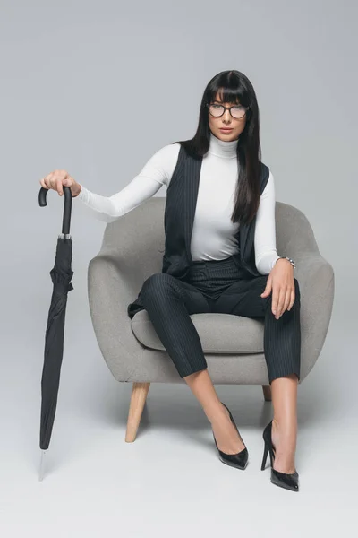 Приваблива брюнетка бізнес-леді сидить на кріслі з парасолькою на сірому — стокове фото
