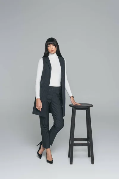 Attraente bruna donna in piedi vicino sedia di legno su grigio — Foto stock