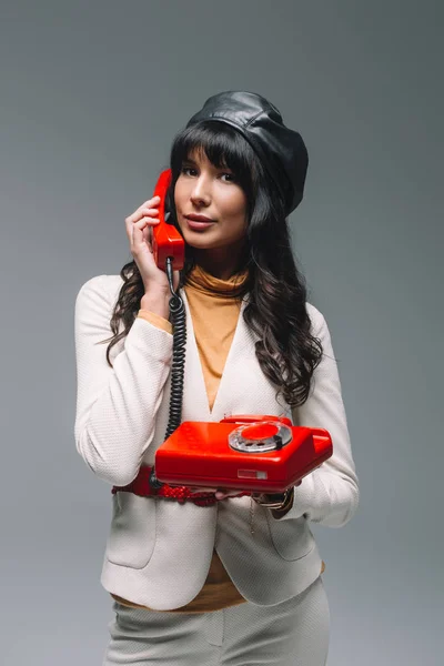 Attractive brunette woman in white suit talking by red landline phone isolated on gray — Stock Photo