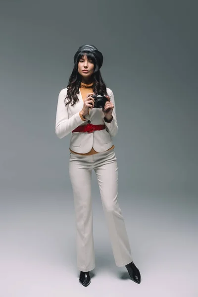 Atractiva mujer morena en traje blanco de pie con cámara de cine en gris - foto de stock