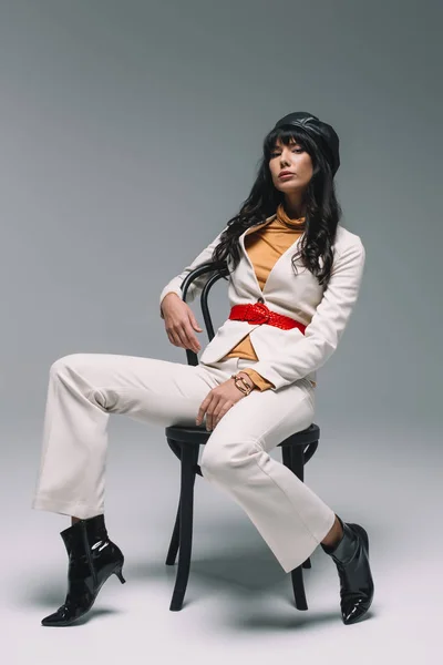 Atractiva mujer morena en traje blanco sentado y posando en silla en gris - foto de stock