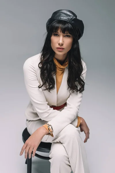 Beautiful brunette woman in white suit sitting on chair and looking at camera isolated on gray — Stock Photo