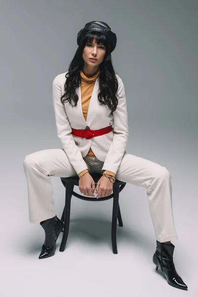 Attractive brunette woman in white suit posing on chair on gray — Stock Photo