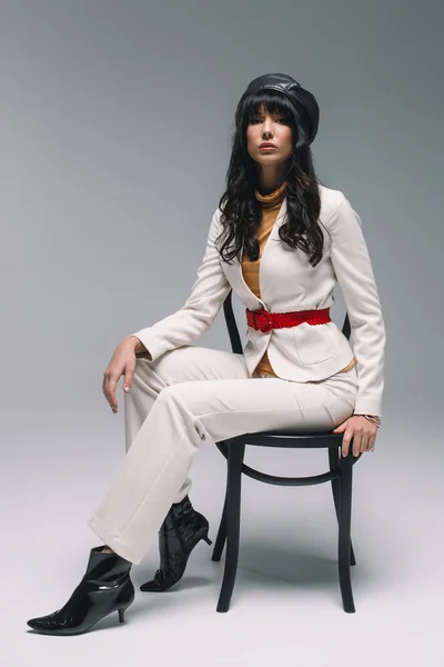 Beautiful brunette woman in white suit sitting on chair on gray — Stock Photo