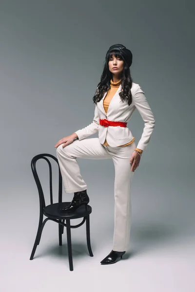 Schöne brünette Frau im weißen Anzug legt Bein auf Stuhl auf grau — Stockfoto