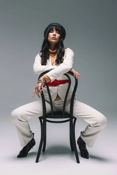 Belle femme brune en costume blanc assise sur une chaise et regardant la caméra sur gris — Photo de stock