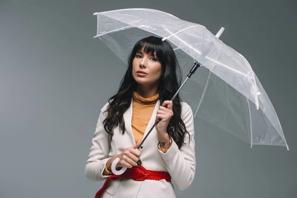 Morena mujer en chaqueta blanca de pie con paraguas aislado en gris - foto de stock