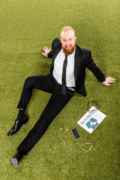 High angle view of bearded businessman smiling at camera while sitting on green grass with smartphone, earphones, eyeglasses and newspaper — Stock Photo