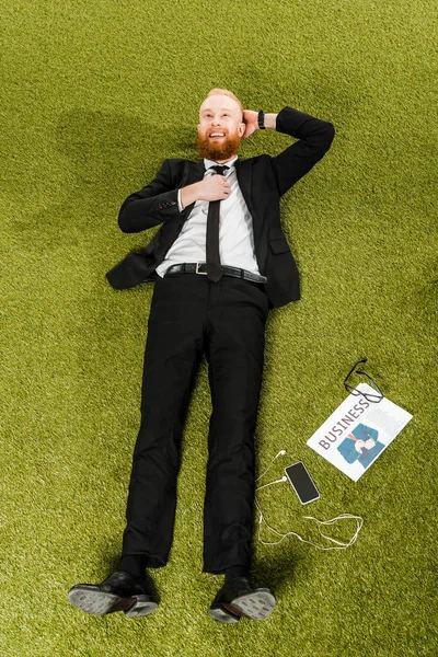 Vista de ángulo alto de sonriente joven hombre de negocios acostado sobre hierba verde con teléfono inteligente, auriculares, gafas y periódico - foto de stock