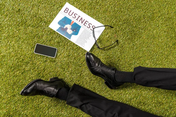 Обрезанный снимок мужских ног в стильной кожаной обуви, очках, газетах и смартфонах с чистым экраном на зеленой траве — стоковое фото