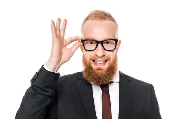 Bearded young businessman adjusting eyeglasses and smiling at camera isolated on white — Stock Photo