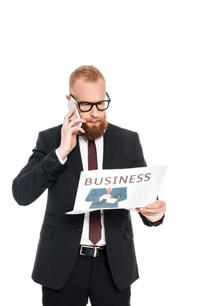 Young bearded businessman in eyeglasses reading newspaper and talking on smartphone isolated on white — Stock Photo