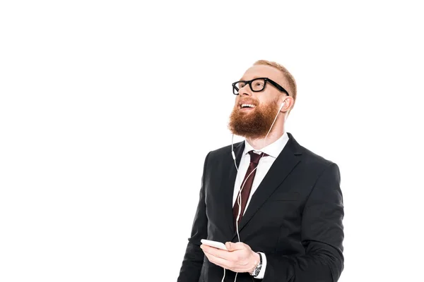 Smiling bearded businessman in earphones listening music with smartphone and looking up isolated on white — Stock Photo