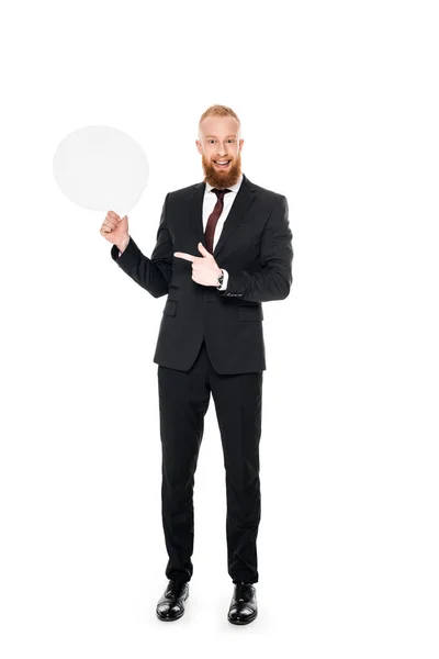 Sonriente hombre de negocios barbudo sosteniendo burbuja de habla en blanco y señalando con el dedo aislado en blanco - foto de stock