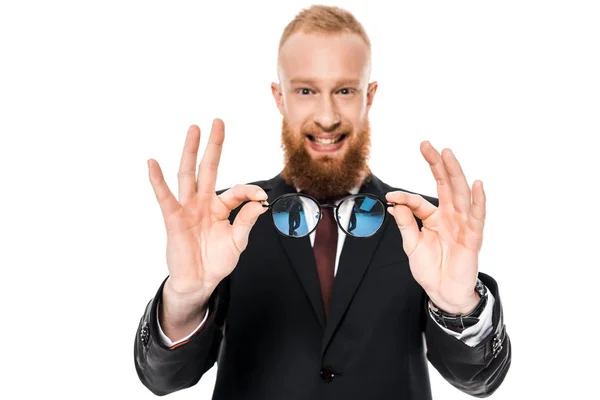 Bearded young businessman holding eyeglasses and smiling at camera isolated on white — Stock Photo