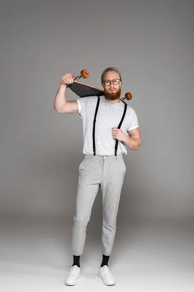 Visão de comprimento total de homem barbudo elegante bonito em óculos segurando longboard e olhando para a câmera em cinza — Fotografia de Stock