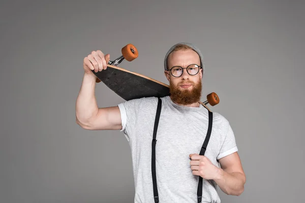 Handsome bearded man holding longboard on shoulders and looking at camera on grey — Stock Photo