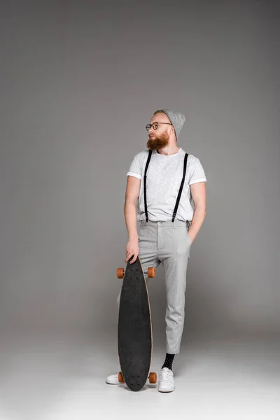 Schöner, stylischer bärtiger Mann, der mit Skateboard steht und wegschaut auf grau — Stockfoto